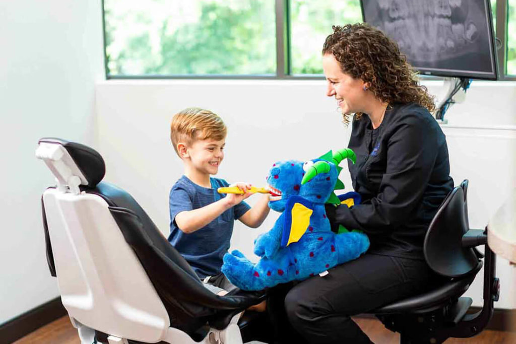 A dental assistant demonstrates good brushing techniques using a stuffed dragon toy to a young dental patient
