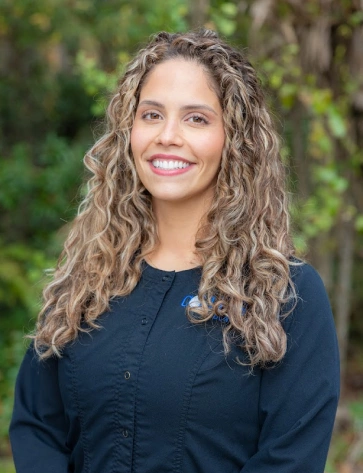 Headshot of a smiling woman with long, curly light-brown and blonde hair standing outdoors. She is wearing a black scrub top with a subtle blue logo, set against a natural green leafy background.
