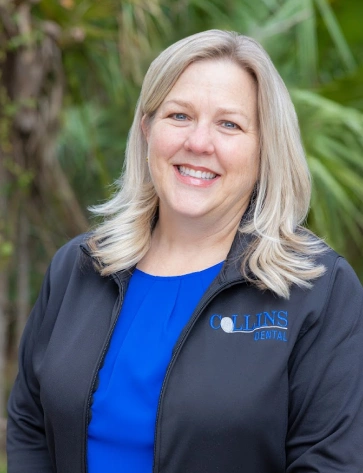 Headshot of a smiling woman with shoulder-length blonde hair standing outdoors. She is wearing a black zip-up jacket with a blue "Collins Dental" logo over a bright blue top, with a green tropical background.