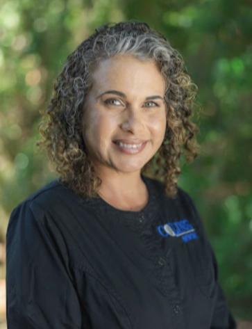 Outdoor headshot of a smiling woman with shoulder-length curly brown hair accented with grey. She is wearing black medical scrubs with a partially visible blue embroidered logo, set against a softly blurred background of green foliage.