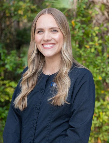 Headshot of a smiling young woman with long, wavy blonde hair. She is wearing a buttoned dark navy or black shirt, standing outdoors in front of a blurred green foliage background.