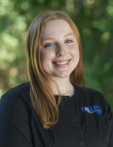 Outdoor headshot of a smiling young woman with straight, light brown hair. She is wearing a black collared shirt or scrub top featuring a blue embroidered logo on the chest, with a blurred green background.