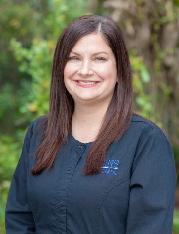Headshot of a smiling woman with long, straight dark brown hair. She is wearing a dark navy or black scrub top featuring a blue embroidered "Collins Dental" logo, set against a softly blurred natural green backdrop.
