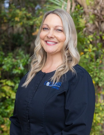 Headshot of a smiling woman with wavy blonde hair standing outdoors. She is wearing black scrubs with a blue embroidered logo on the left chest, positioned against a background of green trees and plants.