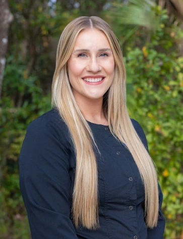 Headshot of a smiling young woman with long, straight blonde hair standing outdoors. She is wearing a buttoned dark navy or black shirt, with a lush green natural backdrop softly out of focus.