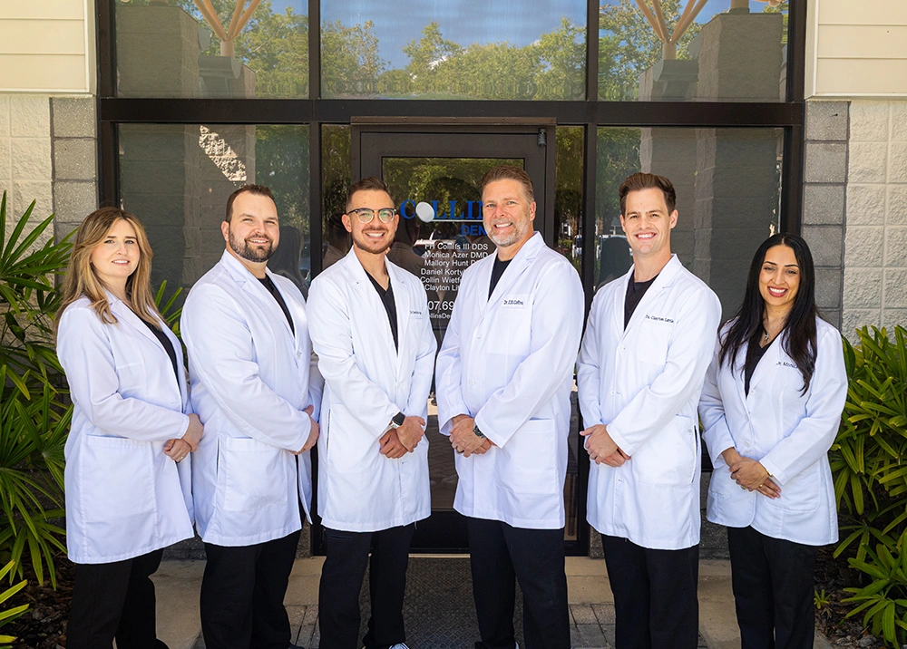 Six dental professionals—two women and four men—stand in a line outside the glass entrance of Collins Dental. They are all wearing white lab coats over dark clothing, smiling at the camera, with green foliage visible on either side of the entrance.