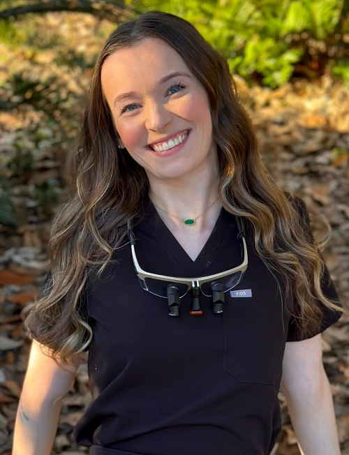 An outdoor portrait of a smiling woman with long brown hair wearing black medical scrubs, a delicate gold necklace with a green stone, and dental loupes resting around her neck.