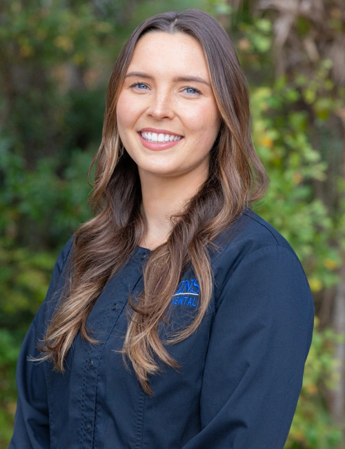 A professional headshot of a smiling young woman with long brown hair, wearing a dark blue button-up scrub shirt, posed against a blurred green foliage background.
