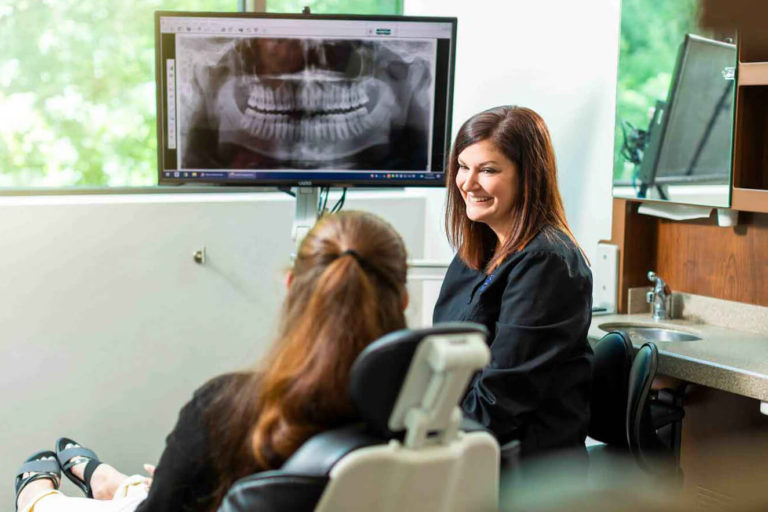 A dental assistant consulting with a female patient who is sitting in an exam chair with a dental x-ray on the screen behind her