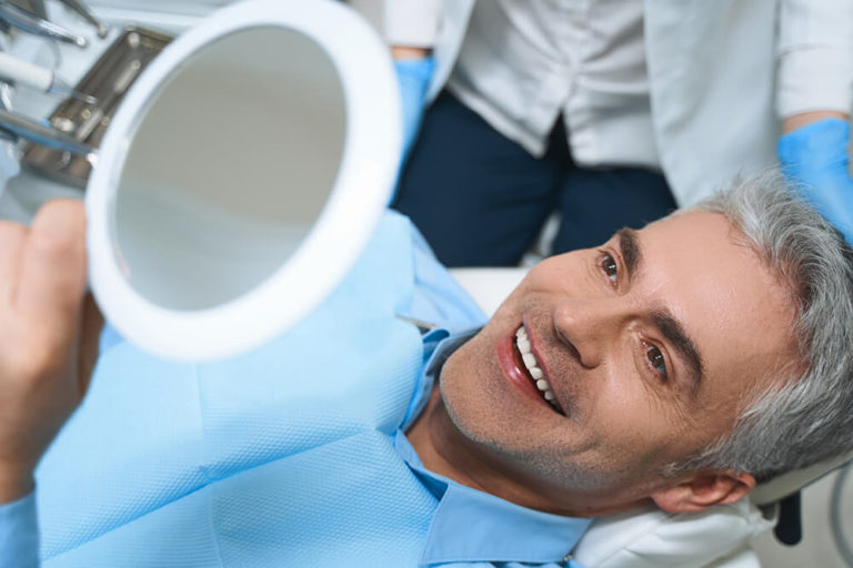 A male dental patient laying back in a dental chair while holding a mirror above his head and smiling as he checks his teeth